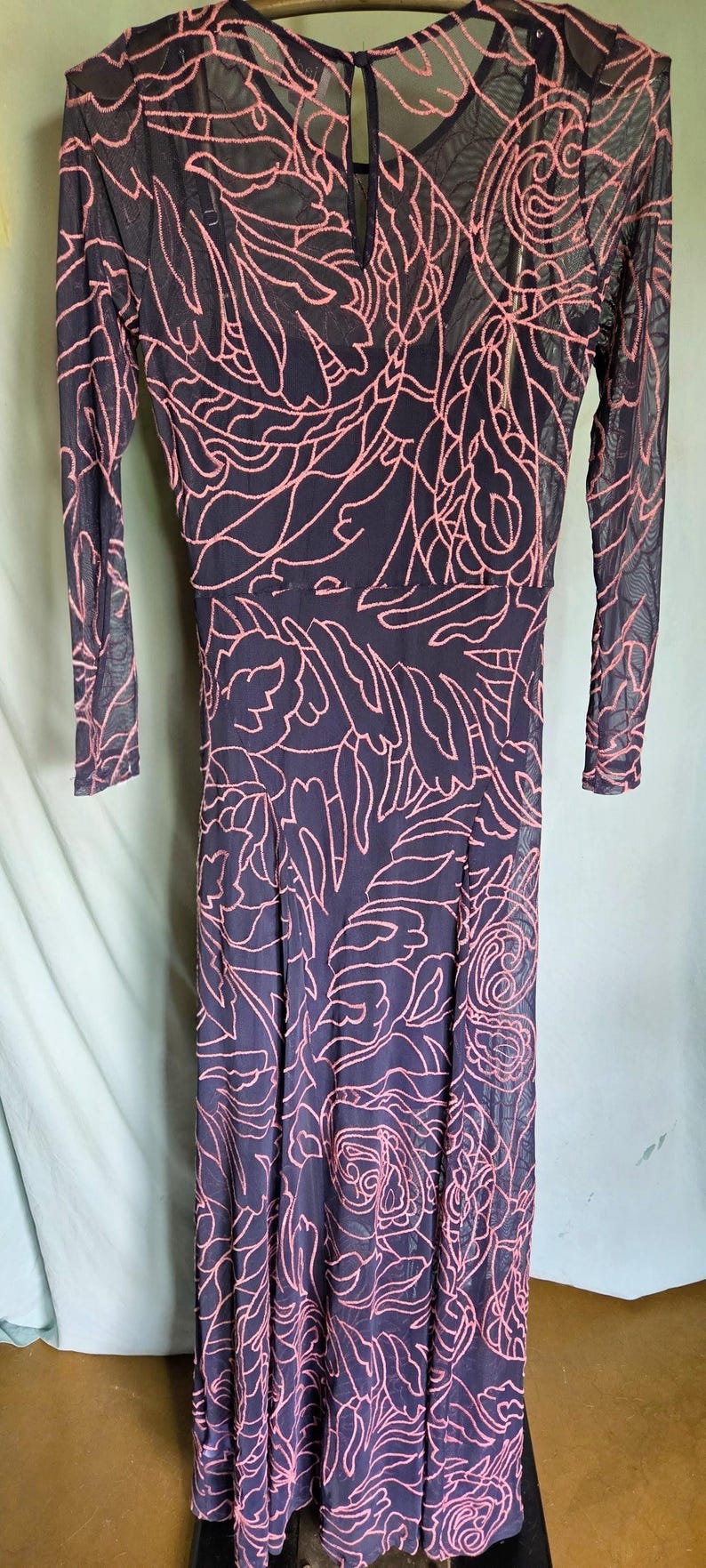 May include: A long, dark blue dress with a pink floral pattern. The dress has long sleeves and a sheer overlay. The floral design is intricate and covers the entire dress. The dress is floor-length and appears to be made of a lightweight material.
