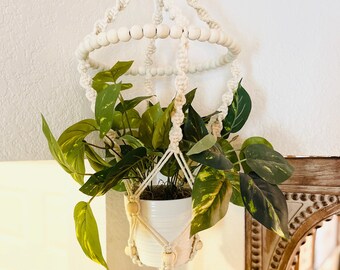 May include: A white macrame plant hanger with wooden beads, holding a white pot with a green plant. The hanger is suspended from a ceiling hook.