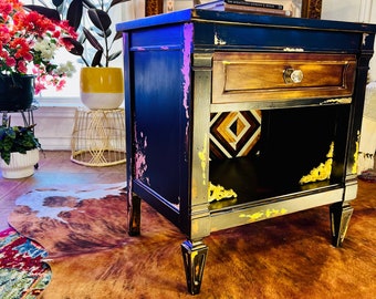 May include: A distressed black nightstand with gold accents and a single drawer. The nightstand has a shelf and is sitting on a cowhide rug.