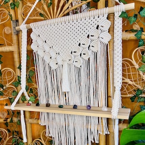 May include: A white macrame wall hanging with a wooden shelf and colorful stones. The macrame is made with a knotting technique and has a fringe at the bottom. The shelf is attached to the macrame with a rope and has a natural wood finish.