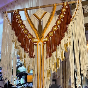 Op de afbeelding: Een groot macramé wandkleed met een geometrisch ontwerp in tinten wit, bruin en geel. Het wandkleed heeft lange franjes en een centrale diamantvorm.