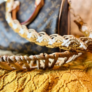 May include: A close-up of a brown leather handbag with a white and brown crocheted strap. The strap is resting on a woven basket.
