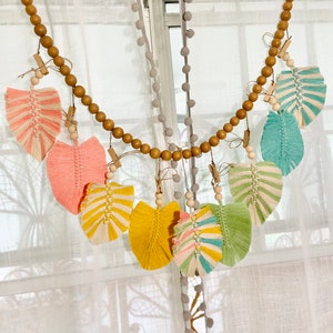 Puede incluir: Una guirnalda de cuentas de madera con borlas de hilo de colores que cuelgan de ella. Las borlas son en tonos de rosa, amarillo, verde y azul. La guirnalda está colgada de una cortina blanca.