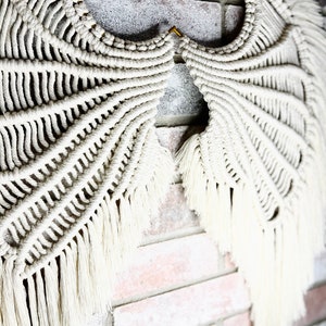 K&ouml;nnte beinhalten: Ein wei&szlig;er Makramee-Wandbehang in Form von Fl&uuml;geln, mit einem goldenen Haken, vor einem Hintergrund aus Backstein.