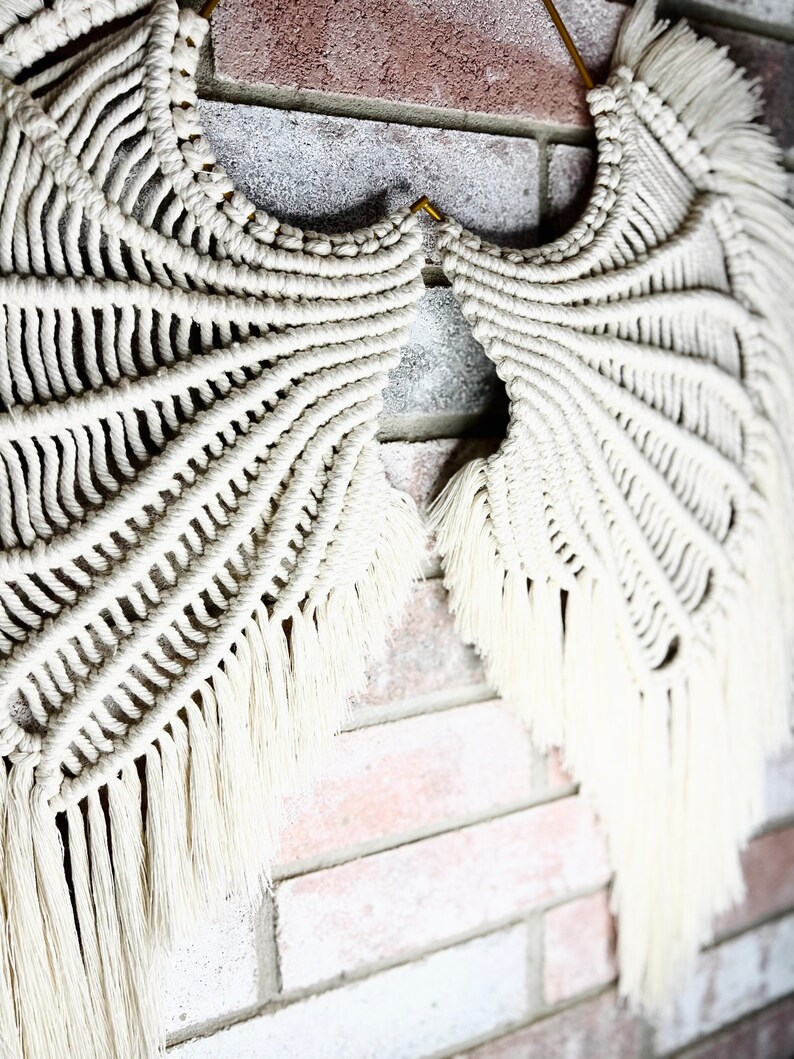 K&ouml;nnte beinhalten: Ein wei&szlig;er Makramee-Wandbehang in Form von Fl&uuml;geln, mit einem goldenen Haken, vor einem Hintergrund aus Backstein.