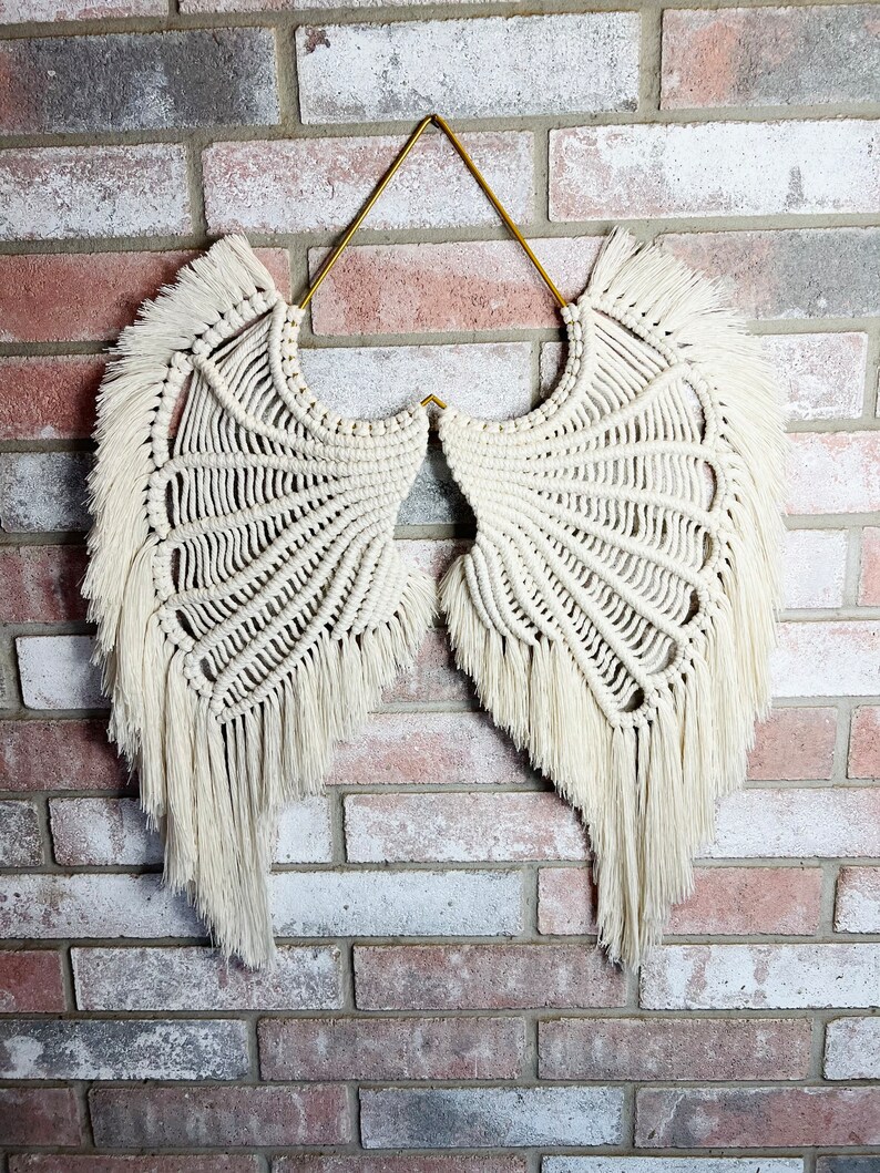 K&ouml;nnte beinhalten: Wei&szlig;e Makramee-Engelsfl&uuml;gel mit einem goldenen Metallhalter. Die Fl&uuml;gel haben einen Fransenbesatz und h&auml;ngen an einer Backsteinwand.