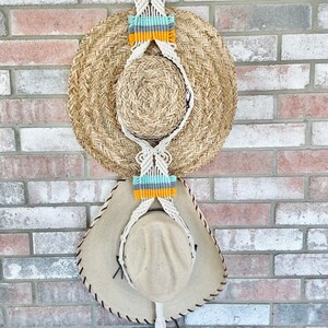 May include: A macrame hat rack with a wooden ring at the top. Two straw hats are hanging from the rack, one on top of the other. The rack is decorated with colorful yarn.