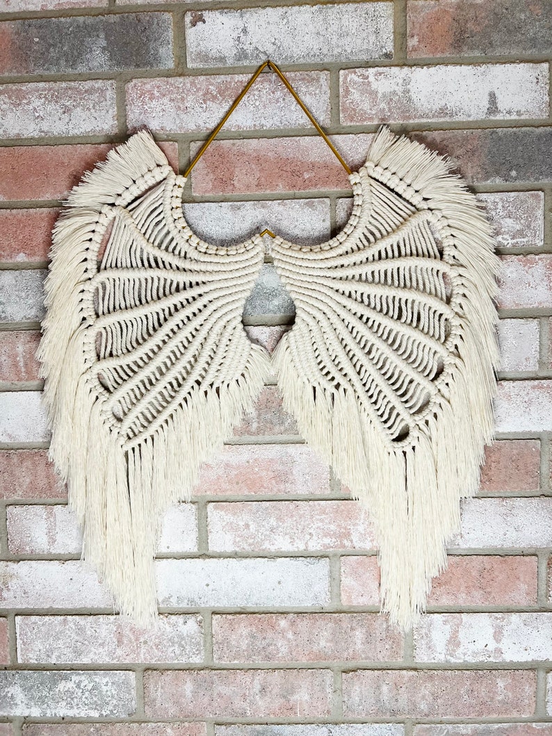 K&ouml;nnte beinhalten: Wei&szlig;er Makramee-Wandbehang in Form von Engelsfl&uuml;geln mit einem goldenen Aufh&auml;nger. Die Fl&uuml;gel haben ein Fransen-Detail.