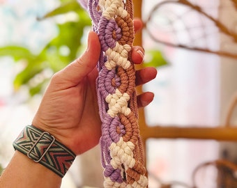 May include: A macrame headband with a white, brown, and purple color scheme. The headband is made with a series of knots and loops.