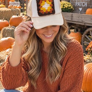 May include: A beige trucker hat featuring a square, multi-colored crochet granny square patch on the front panel. The patch displays a geometric pattern in shades of white, yellow, orange, and dark brown, stitched onto the solid beige fabric.