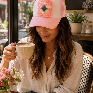 May include: A pink and white trucker hat with a crocheted square patch. The patch features a pink and cream-colored floral design with a dark gray center. The hat has a pink brim and a white front panel.