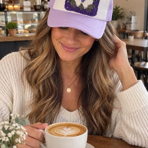 May include: A lavender and white trucker hat featuring a square crochet patch on the front panel. The patch displays a geometric pattern in shades of purple, dark gray, and white. The hat has a solid lavender brim and a white mesh back section.