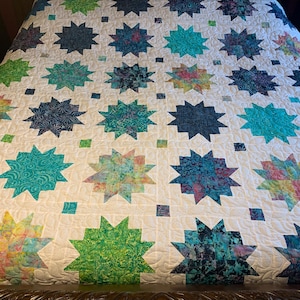May include: A white quilt with a repeating pattern of starbursts in shades of blue, green, and yellow. The quilt is made of fabric and has a quilted texture. The starbursts are arranged in rows and columns.