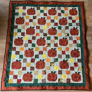 May include: A quilted blanket featuring a repeating pattern of detailed, textured pumpkins in shades of orange and brown. The pumpkins are set against a cream background with colorful square accents. The quilt has a green and rust-colored border.