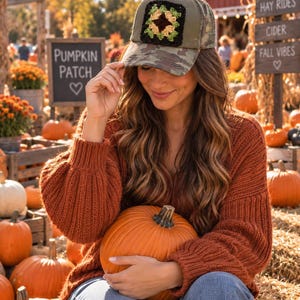 May include: An olive green camouflage baseball cap featuring a square crochet patch on the front. The patch displays a geometric pattern in shades of black, green, and tan. The cap has a structured front panel and a camouflage brim.