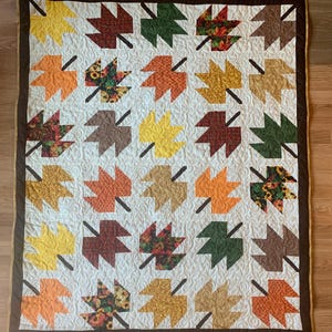 May include: A quilted blanket featuring a repeating pattern of stylized autumn leaves in shades of yellow, orange, brown, and green against a white background. The leaves have black stems and are bordered by a brown trim.
