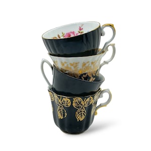 May include: Four stacked teacups with ornate designs. The cups are black, white, and gold, with floral and leaf patterns. One cup features a pink rose design. Each cup has a handle and a gold trim.