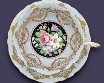 May include: A blue and gold teacup and saucer with a floral design. The cup features a black background with a pink, white, and purple floral pattern. The saucer has a gold rim and a delicate floral pattern.