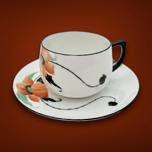 May include: A white porcelain teacup and saucer with a black rim and floral design. The design features orange poppy flowers with black stems and leaves.