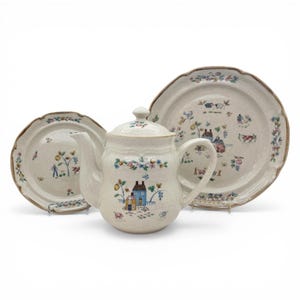 Puede incluir: Un juego de té de cerámica color crema con una tetera y dos platos. El juego está decorado con una escena rural de casas, árboles y flores. Los platos y la tetera tienen un borde festoneado con un ribete marrón.