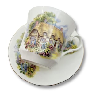 May include: White teacup and saucer set with a cottage scene. The teacup and saucer are white with a gold trim. The teacup features a detailed illustration of a thatched-roof cottage surrounded by colorful flowers and trees. The saucer has a matching design.