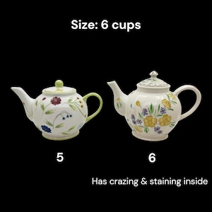 May include: Two white ceramic teapots with floral designs. One teapot has a green handle and lid, the other has a white handle and lid. The text "Size: 6 cups" is above the teapots. The text "Has crazing & staining inside" is below the teapots.
