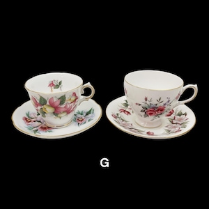 May include: Two white teacups and saucers with floral designs and gold trim. One cup features pink and yellow flowers, the other has red roses. Both are on a black background.