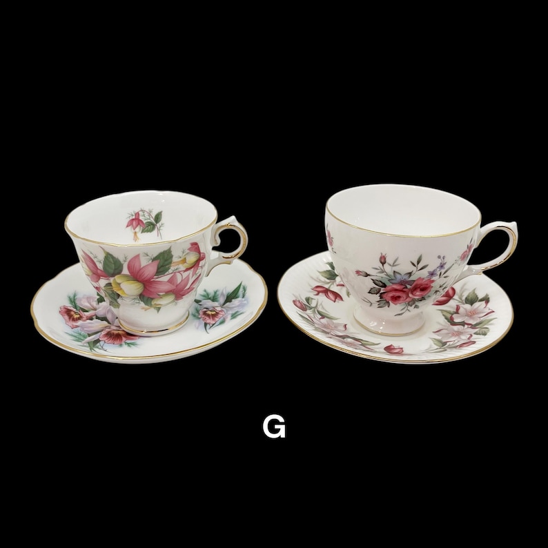 May include: Two white teacups and saucers with floral designs and gold trim. One cup features pink and yellow flowers, the other has red roses. Both are on a black background.
