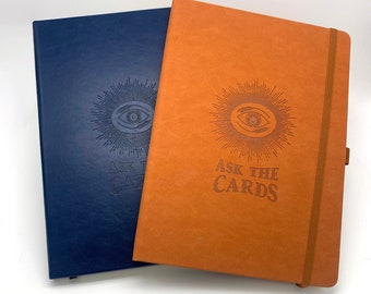 Può includere: Due diari rilegati con copertina blu e arancione. Entrambi i diari presentano un occhio in rilievo con un motivo a raggiera e il testo "Ask the Cards".