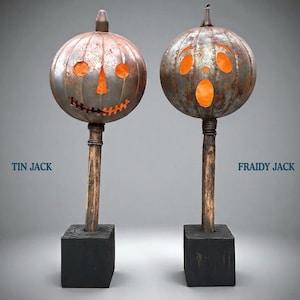 May include: Two metal pumpkin sculptures on wooden posts and black square bases. The left sculpture, labelled TIN JACK, has a stitched mouth and triangular eyes. The right sculpture, labelled FRAIDY JACK, has wide eyes and an open mouth.