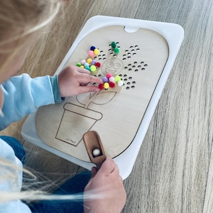 Op de afbeelding: Een houten bord met een bloemetjesontwerp en gaten voor pompoenen. Een kind speelt met het speelgoed en plaatst kleurrijke pompoenen in de gaten. Het bord heeft een witte rand en een kleine houten schep om de pompoenen op te scheppen.