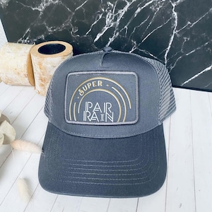 Peut inclure: Casquette de camionneur grise avec un dos en filet et une visière incurvée. Le panneau avant présente un écusson rectangulaire avec les mots "SUPER PAIR RAIN" en lettres dorées et blanches. Idéale pour un usage décontracté.