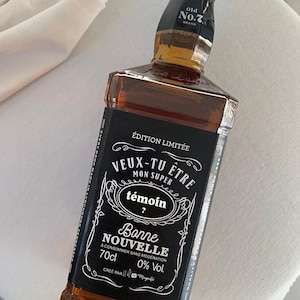May include: A brown glass bottle with a black label featuring white text. The label includes the words "VEUX-TU ETRE MON SUPER témoin?" and "Bonne NOUVELLE". The bottle contains a dark amber liquid and is labeled "70cl".