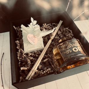 Peut inclure: Une boîte cadeau noire remplie de papier déchiqueté brun, un savon blanc avec le texte "veux-tu etre ma marraine" en rose, une bouteille en verre d'huile de diffuseur avec le texte "Marraine en Or" en or, et un ensemble de bâtonnets de diffuseur en bois.