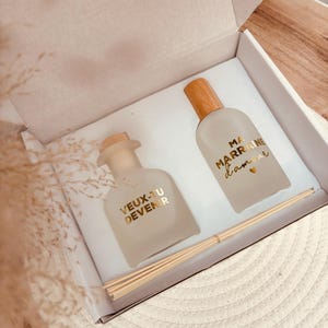 May include: A gift set with two glass bottles with wooden tops and gold labels. The labels read "Veux-tu devenir" and "Ma marraine d'amour". The set includes a bundle of wooden diffuser sticks.