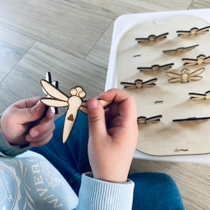 May include: A child's hand holds a wooden dragonfly toy. The toy is light brown with black details. The child is playing with a wooden dragonfly matching game. The game board has multiple slots for the dragonfly pieces.