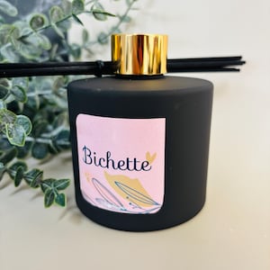 Op de afbeelding: Een zwarte diffuserfles met een gouden dop en zwarte stokjes. De fles heeft een roze label met het woord "Bichette" in sierlijke letters en bloemenillustraties. De diffuser is voor huisgeur.
