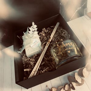 Könnte beinhalten: Eine schwarze Geschenkbox gefüllt mit braunem Packpapier. Die Box enthält eine weiße Seife mit einem Etikett mit der Aufschrift "Lavender &  Ground-nut Oil", eine Glasflasche mit Diffuseröl mit einem goldenen Etikett mit der Aufschrift "MAMY  Amour" und einen Satz Holzdiffuserstäbchen.