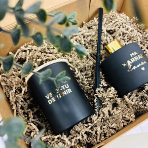 Könnte beinhalten: Ein schwarzes Kerzen- und Diffusor-Set mit goldfarbenen Schriftzügen in einer Geschenkbox. Auf der Kerze steht "VEUX-TU DEVENIR" und auf dem Diffusor "MA MARRAINE D'AMOUR". Das Set enthält einen schwarzen Diffusor-Stick.