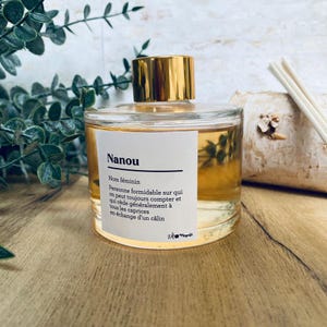 Può includere: Un flacone di profumo in vetro trasparente con tappo dorato ed etichetta bianca con la scritta "Nanou". Il flacone contiene un liquido ambrato chiaro. L'etichetta include anche testo francese. Il flacone è su una superficie di legno.