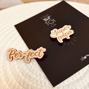 May include: Two wooden pins with the text "Super Papa" and "Per-fect" in a cursive font. The pins are on a black background with the text "C'est tout toi ca" in a white font.