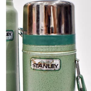 May include: A vintage green Stanley thermos with a textured finish and a shiny silver metal cap. The front features a rectangular label with the text Aladdin STANLEY. A green plastic carrying handle is attached to the side of the cylindrical body.