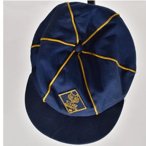 May include: A navy blue cap with yellow piping along the seams and a central button. A diamond-shaped patch on the front features a yellow embroidered wolf head logo and the text CUB SCOUTS. The cap has a rounded crown and a stiff, curved brim.