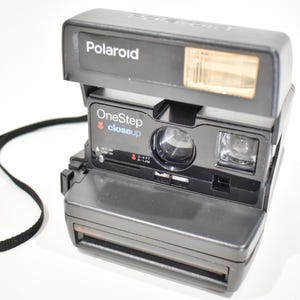 May include: A black Polaroid OneStep closeup instant camera featuring a built-in flash, a lens, and a viewfinder. The front of the camera displays the text Polaroid, OneStep, and closeup, along with a focus range indicator of 2-4 FT and .6-1.2M.