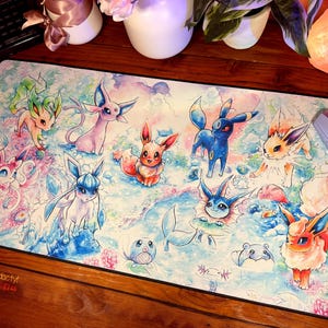 May include: A white mouse pad with a watercolor design featuring various Pokemon characters, including Eevee and its evolutions. The background is a light blue with white clouds and snowflakes. The mouse pad is surrounded by a black border.