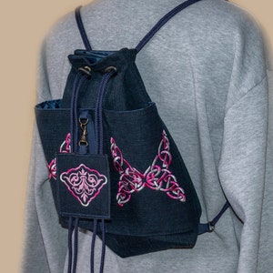 Peut inclure: Un sac à dos bleu foncé à cordon avec une broderie rose et blanche d'un motif de nœud celtique. Le sac à dos a une petite poche zippée à l'avant.