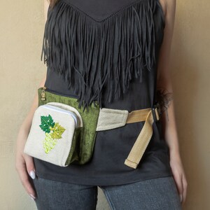 May include: A black fringed top with a green and beige fanny pack featuring an embroidered white grape design.
