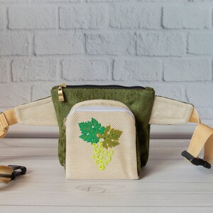 May include: A green and beige fanny pack with a zippered pouch featuring an embroidered grape design. The pouch is attached to the fanny pack with a zipper.