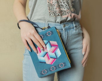 May include: A blue and pink geometric patterned clutch with a gold zipper and a black strap. The clutch is held by a person wearing a grey tank top with a pink and black animal print and blue jeans.