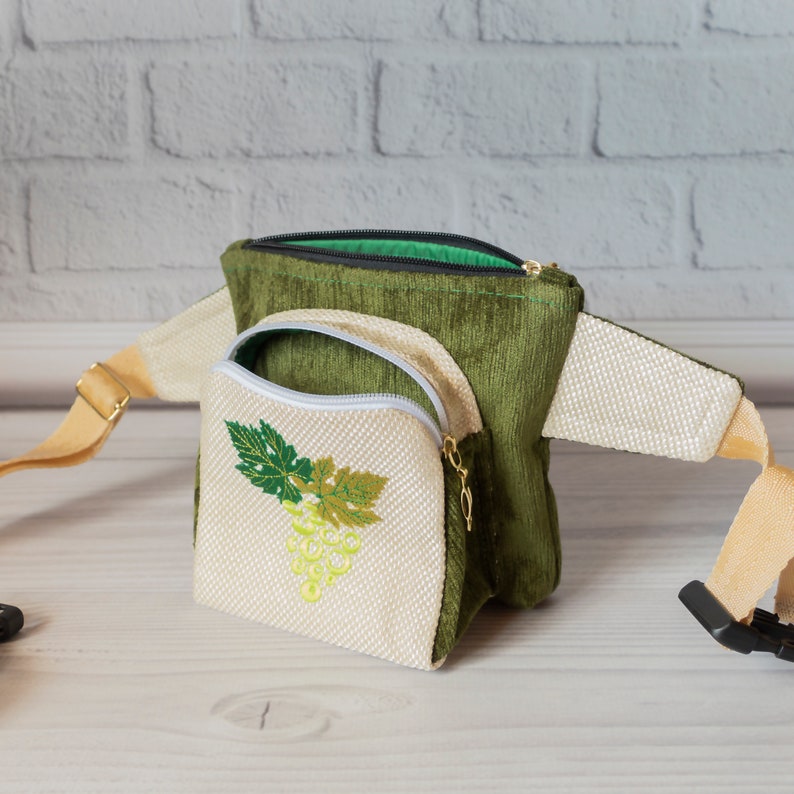May include: A green and cream fanny pack with a zippered pocket featuring an embroidered grape design. The bag has a long adjustable strap.